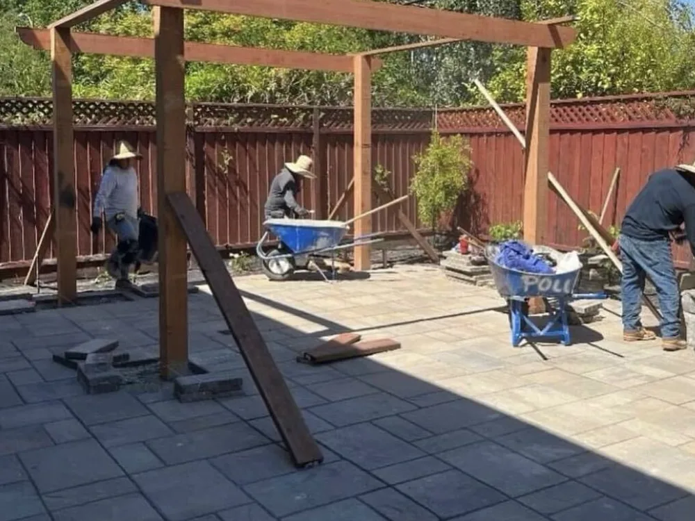 Crew in sun hats raising a redwood pergola frame over a finished paver patio